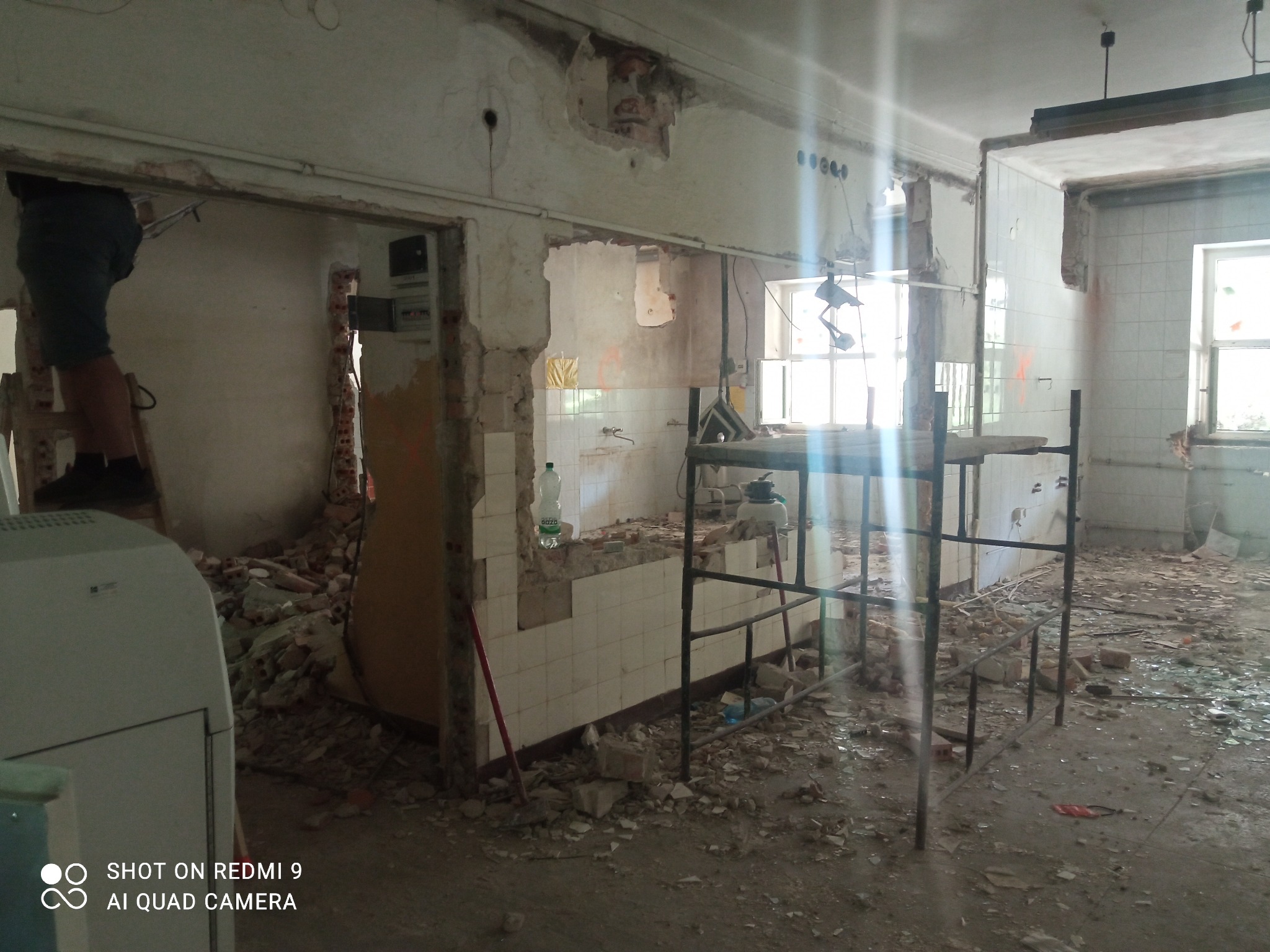Wnętrze zniszczonego pomieszczenia w trakcie rozbiórki, widoczne resztki płytek, gruz, instalacje oraz fragment drabiny i osoba w górnej części kadru.