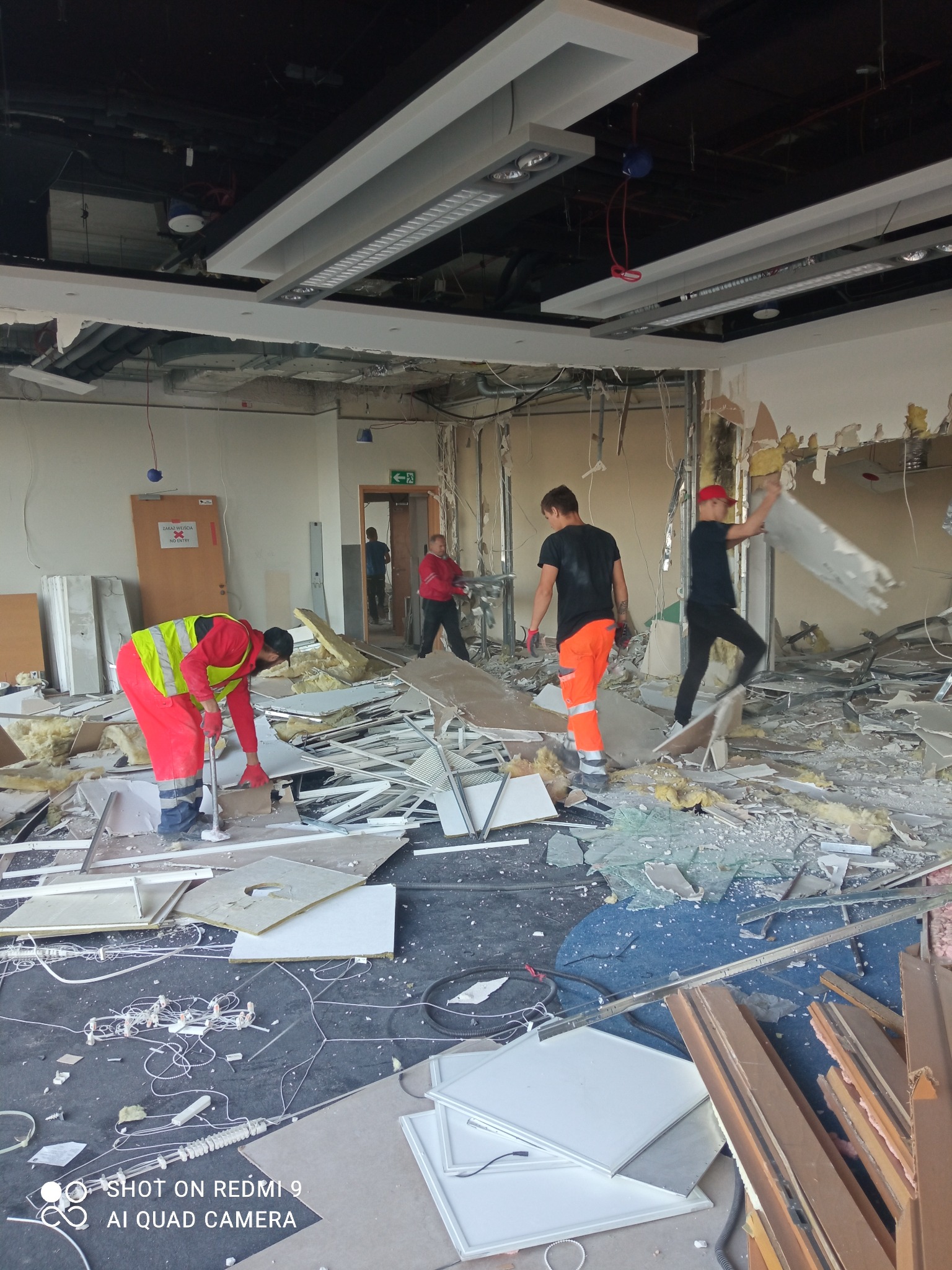 Ekipa w odblaskowych strojach usuwa gruz i resztki zdemontowanego sufitu podwieszanego w trakcie rozbiórki pomieszczenia biurowego. Widoczne kable, fragmenty ścian i ościeżnic.
