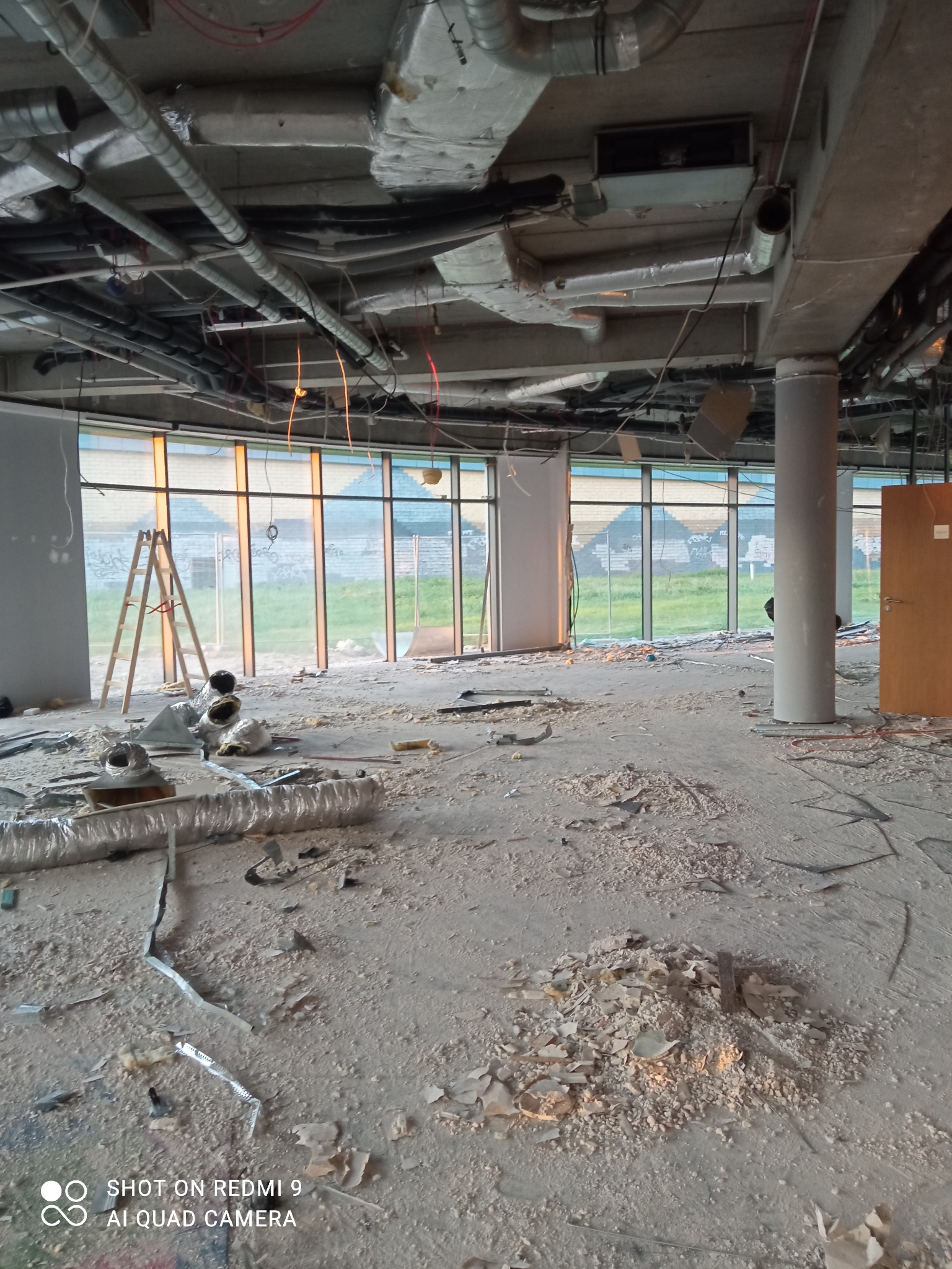 Wnętrze biura w trakcie rozbiórki, widoczne odsłonięte instalacje sufitowe, gruz na podłodze, drabina oparta o okno, kolumna nośna i drzwi w tle.