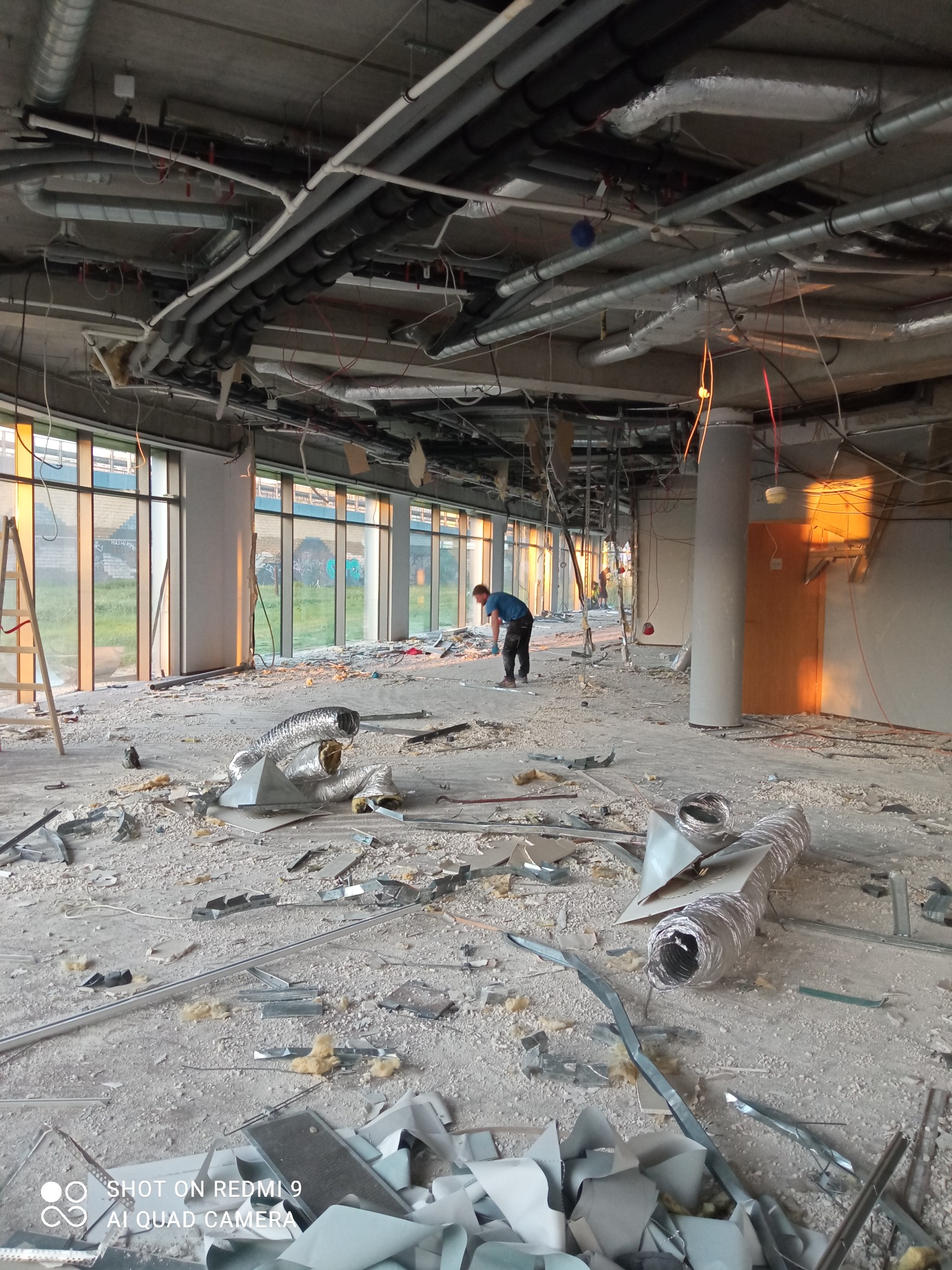 Wnętrze budynku w trakcie rozbiórki, widoczne gruz, fragmenty instalacji, pracownik w oddali oraz rury wentylacyjne na podłodze.