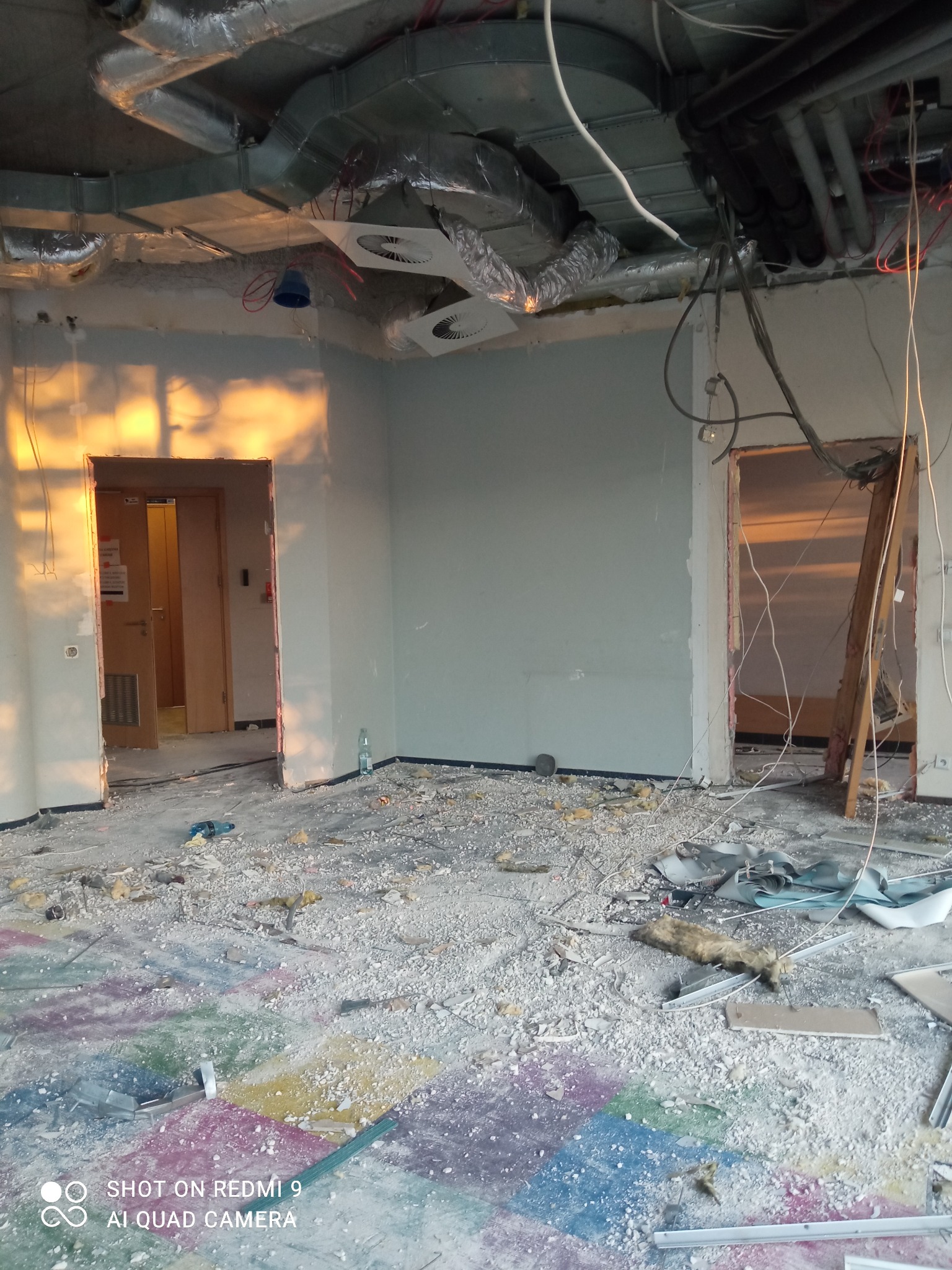Wnętrze pomieszczenia w trakcie rozbiórki, widoczne odsłonięte instalacje pod sufitem, gruz na podłodze z fragmentami kolorowej farby, oraz dwa otwarte przejścia w ścianach.