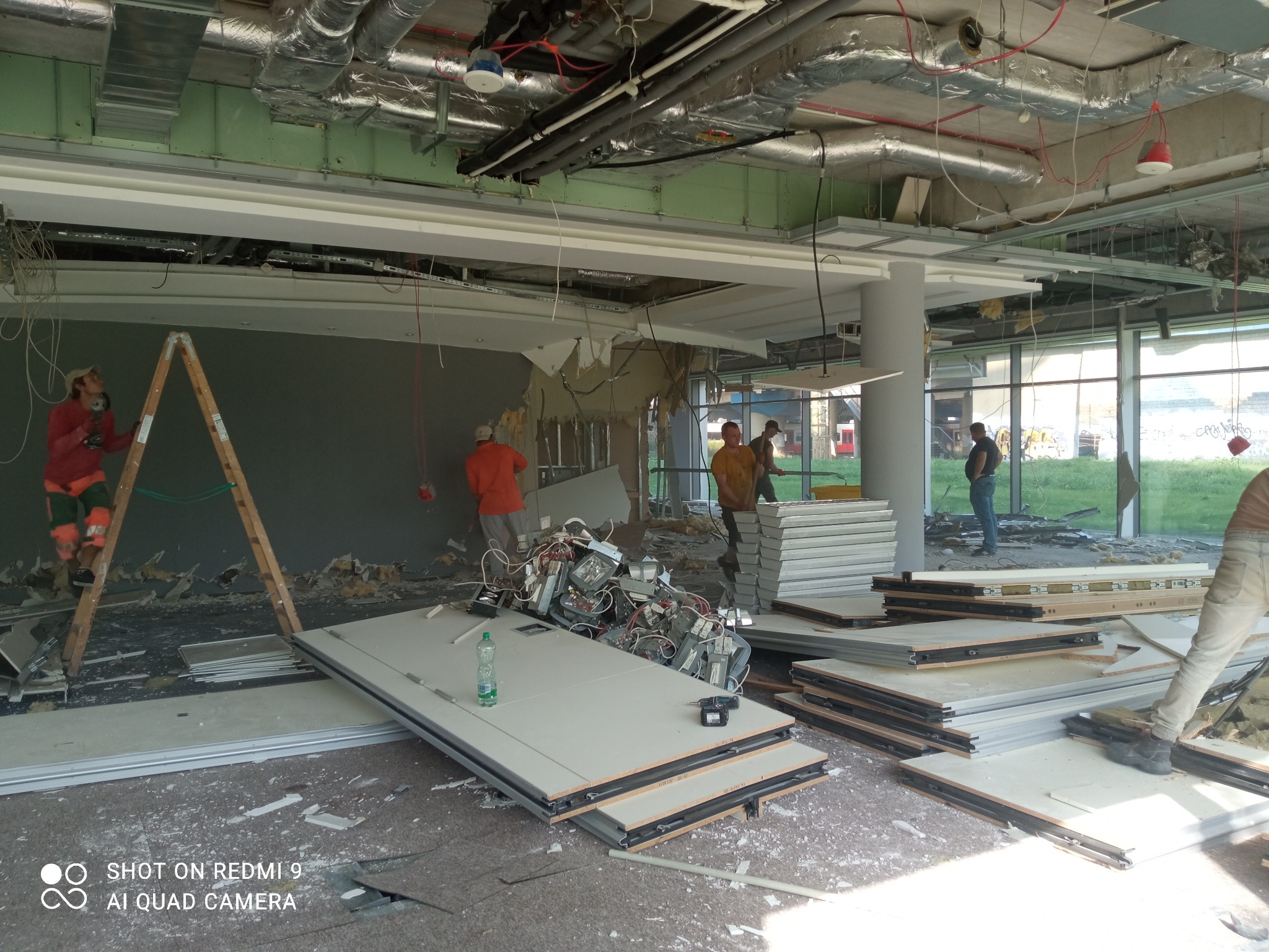 Wnętrze biura w trakcie demontażu ścian działowych, widoczne elementy konstrukcyjne sufitu, pracownicy w odzieży roboczej, drabina oraz stosy zdemontowanych paneli ściennych i oświetlenia.