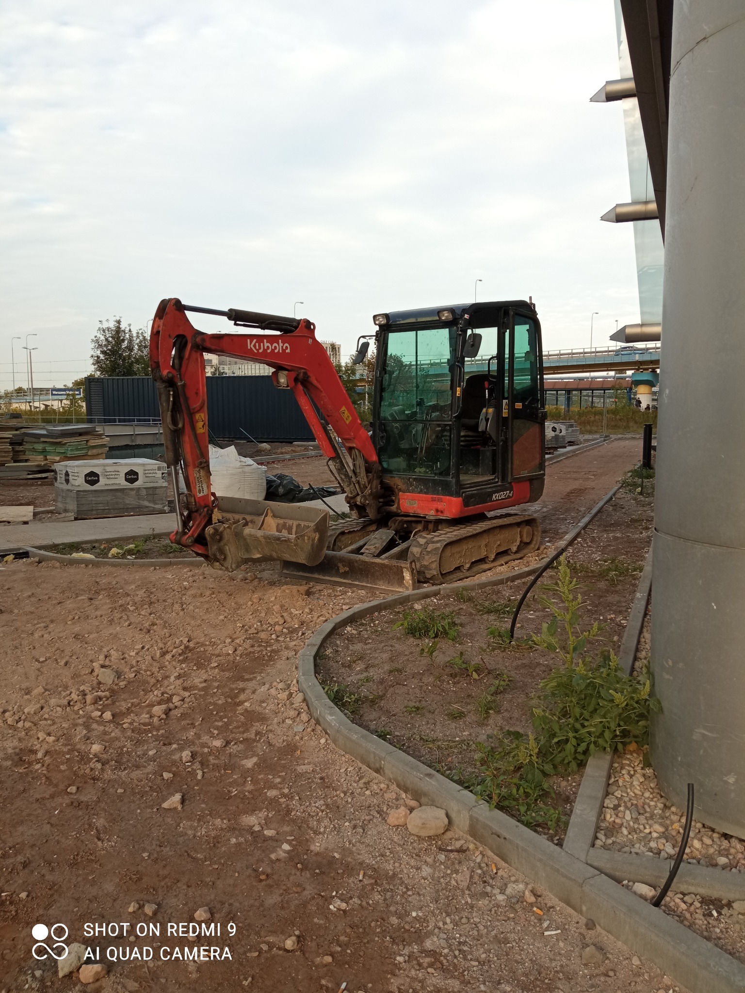 Czerwono-czarna minikoparka Kubota KX027-4 na placu budowy z gruzem i materiałami budowlanymi w tle, widoczny fragment mostu i słup konstrukcyjny.
