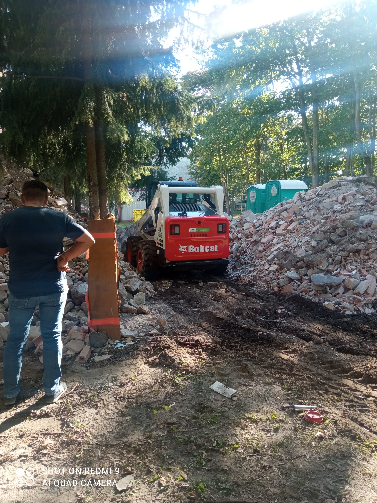 Bobcat na terenie rozbiórki, z stertą gruzu i przenośnymi toaletami w tle, mężczyzna obserwuje prace.