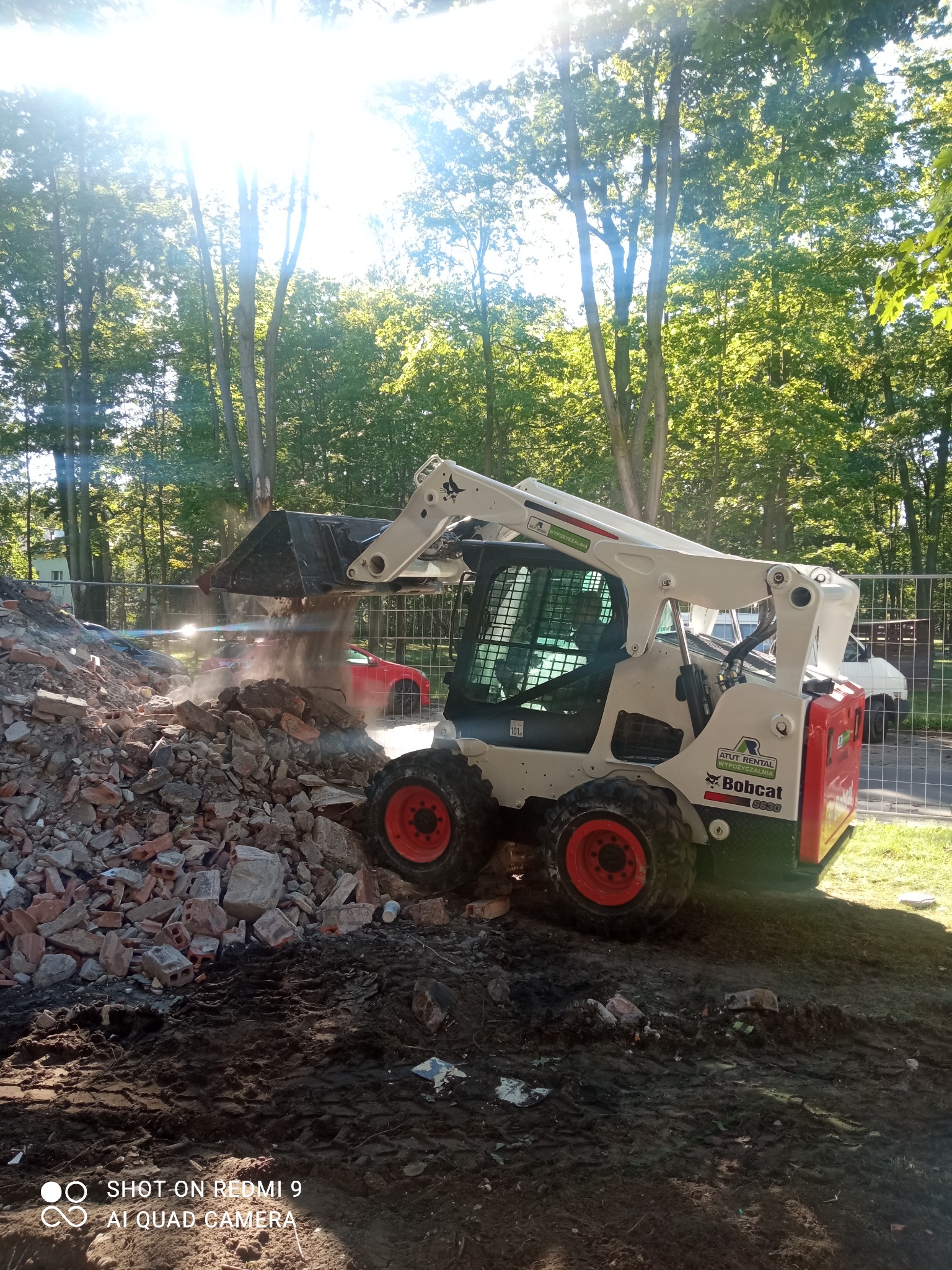 Bobcat S630 z podniesioną łyżką wysypuje gruz ceglany na pryzmę, widoczne ślady opon na ziemi, w tle ogrodzenie, drzewa i częściowo widoczny czerwony samochód.