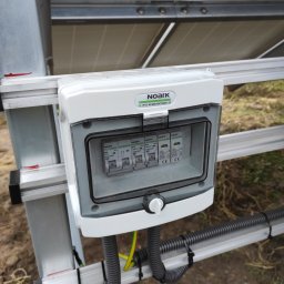 PROMOSOLAR Paweł Gramburg - Szczegółowe ujęcie skrzynki z bezpiecznikami firmy Noark, zamontowanej na konstrukcji wsporczej paneli słonecznych, z widocznymi przewodami w peszlach i uziemieniem.