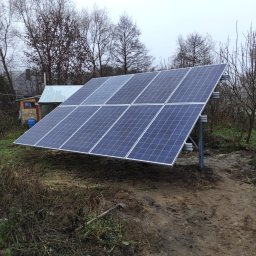 PROMOSOLAR Paweł Gramburg - Zestaw paneli słonecznych zamontowany na metalowej konstrukcji na zewnątrz, widoczne drzewa bez liści i mały budynek w tle, pochmurny dzień.