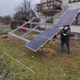 PROMOSOLAR Paweł Gramburg - Montaż paneli słonecznych na metalowej konstrukcji w ogrodzie przy domu z balkonem, widoczne przewody elektryczne i narzędzia.
