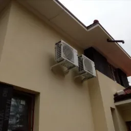 Dwa białe klimatyzatory Mitsubishi Electric zamontowane na beżowej ścianie budynku jednorodzinnego z widocznym oknem i fragmentem dachu z czerwoną dachówką.