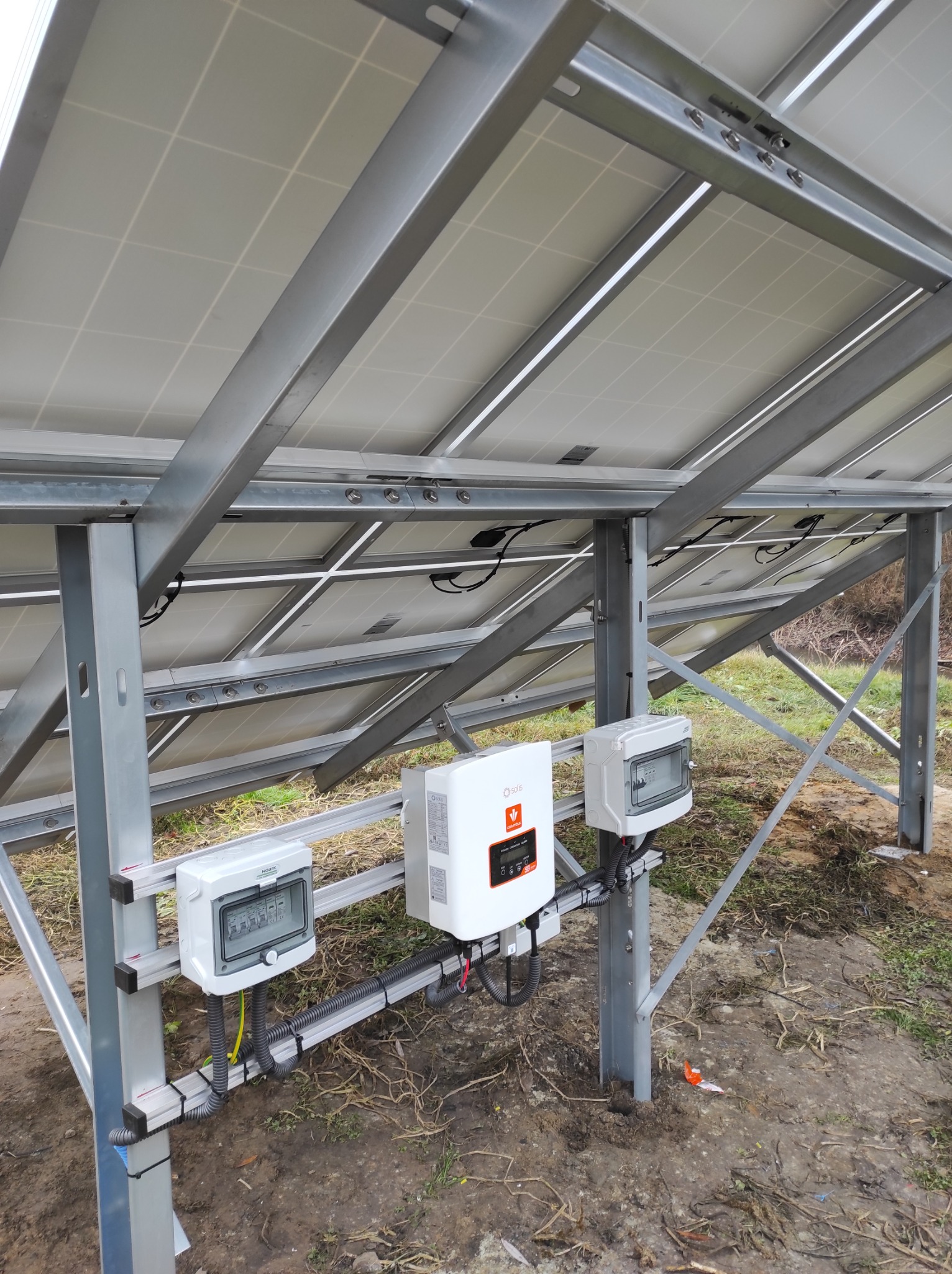 Montaż paneli słonecznych z widocznym inwerterem i zabezpieczeniami elektrycznymi w Serocku, widok od spodu konstrukcji wsporczej.