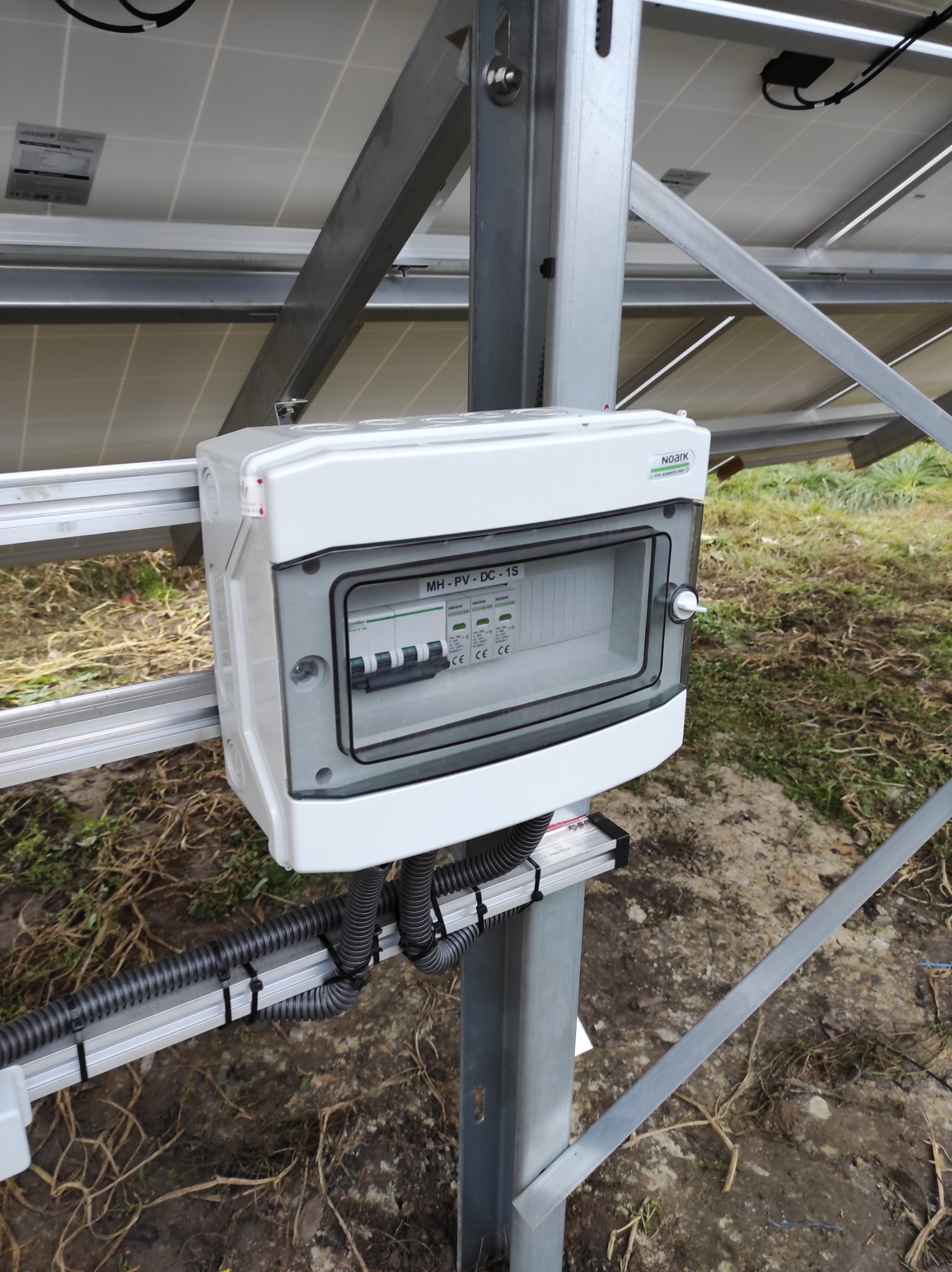 Biała skrzynka zabezpieczeń prądu stałego MH-PV-DC-1S z widocznymi bezpiecznikami, zamontowana na konstrukcji wsporczej paneli słonecznych, z przewodami w peszlach przymocowanych opaskami zaciskowymi.