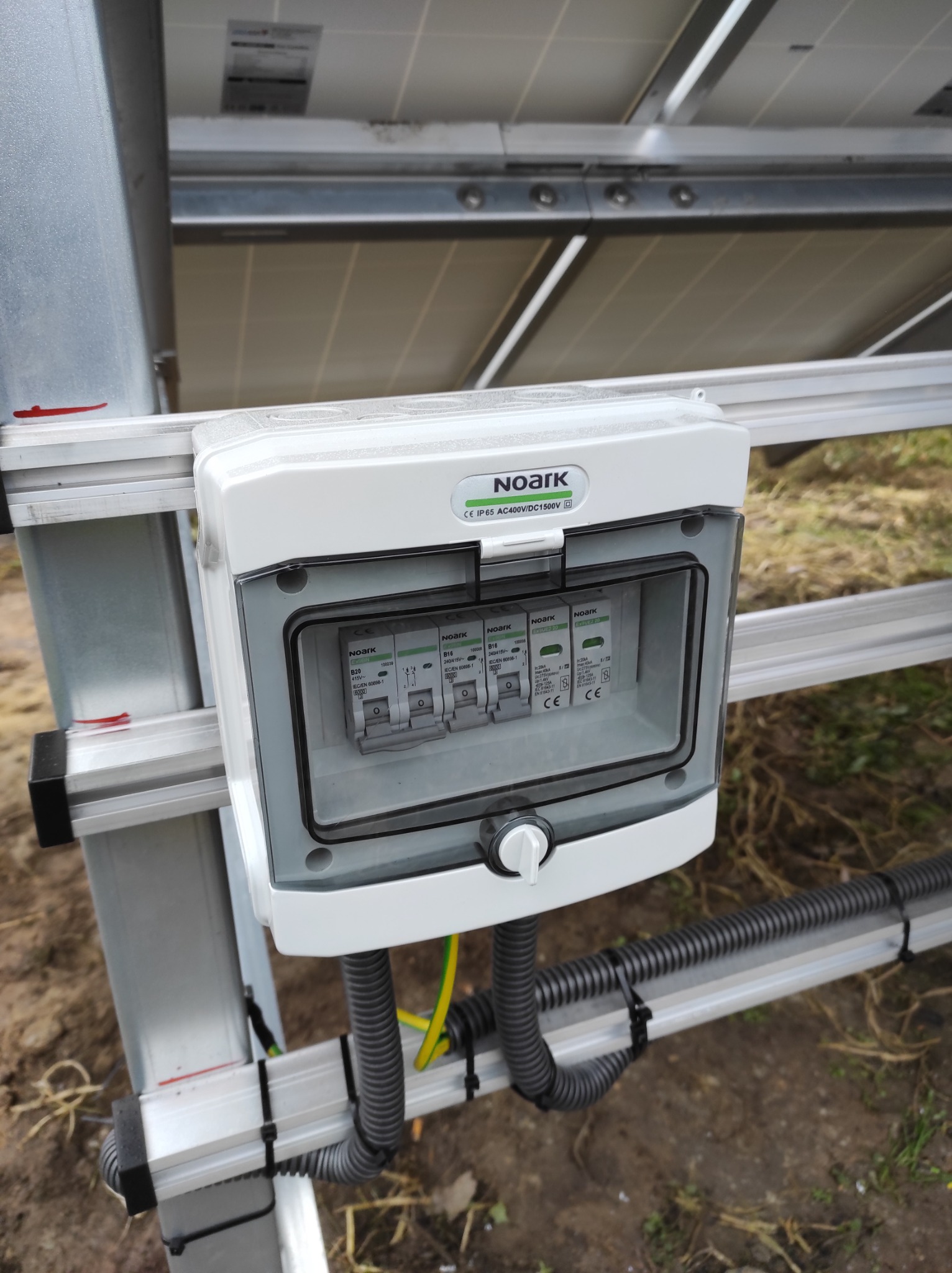 Szczegółowe ujęcie skrzynki z bezpiecznikami firmy Noark, zamontowanej na konstrukcji wsporczej paneli słonecznych, z widocznymi przewodami w peszlach i uziemieniem.
