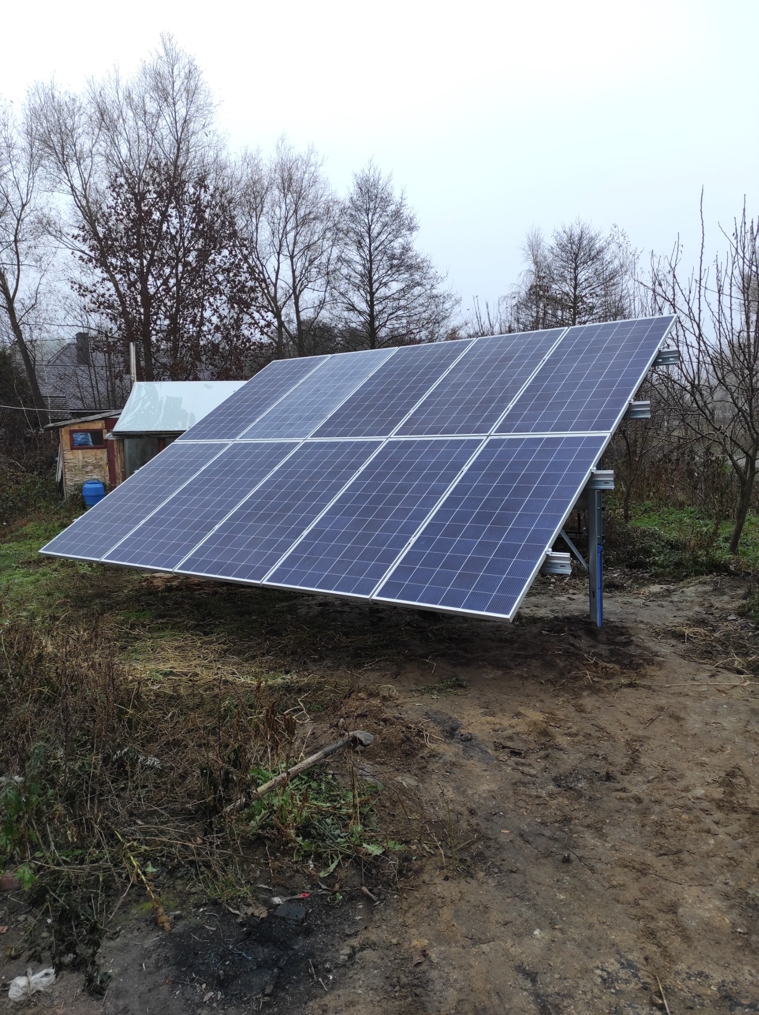 Zestaw paneli słonecznych zamontowany na metalowej konstrukcji na zewnątrz, widoczne drzewa bez liści i mały budynek w tle, pochmurny dzień.