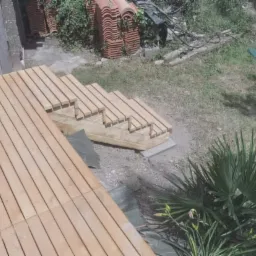 Drewniany taras i schody w ogrodzie, wykonane z jasnych desek, widok z góry, w tle sterta dachówek i zieleń.