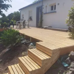 Drewniany taras z schodami prowadzącymi do ogrodu, widok na biały dom z niebieskimi akcentami, meble ogrodowe i rośliny w tle.