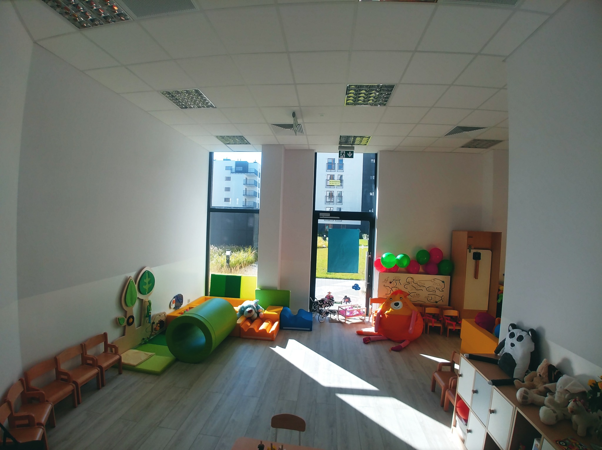 Jasne, przestronne wnętrze biura zaadaptowane na salę zabaw dla dzieci, z elementami wyposażenia w postaci kolorowych mebli, pluszaków i tunelu, oświetlone lampami sufitowymi.