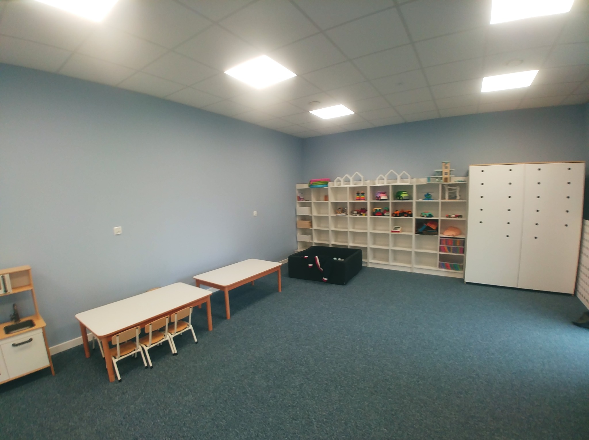 Jasne, przestronne wnętrze pokoju zabaw z niebieskimi ścianami, wyposażone w białe regały z zabawkami, stoliki z krzesłami dla dzieci oraz imitację kuchenki.