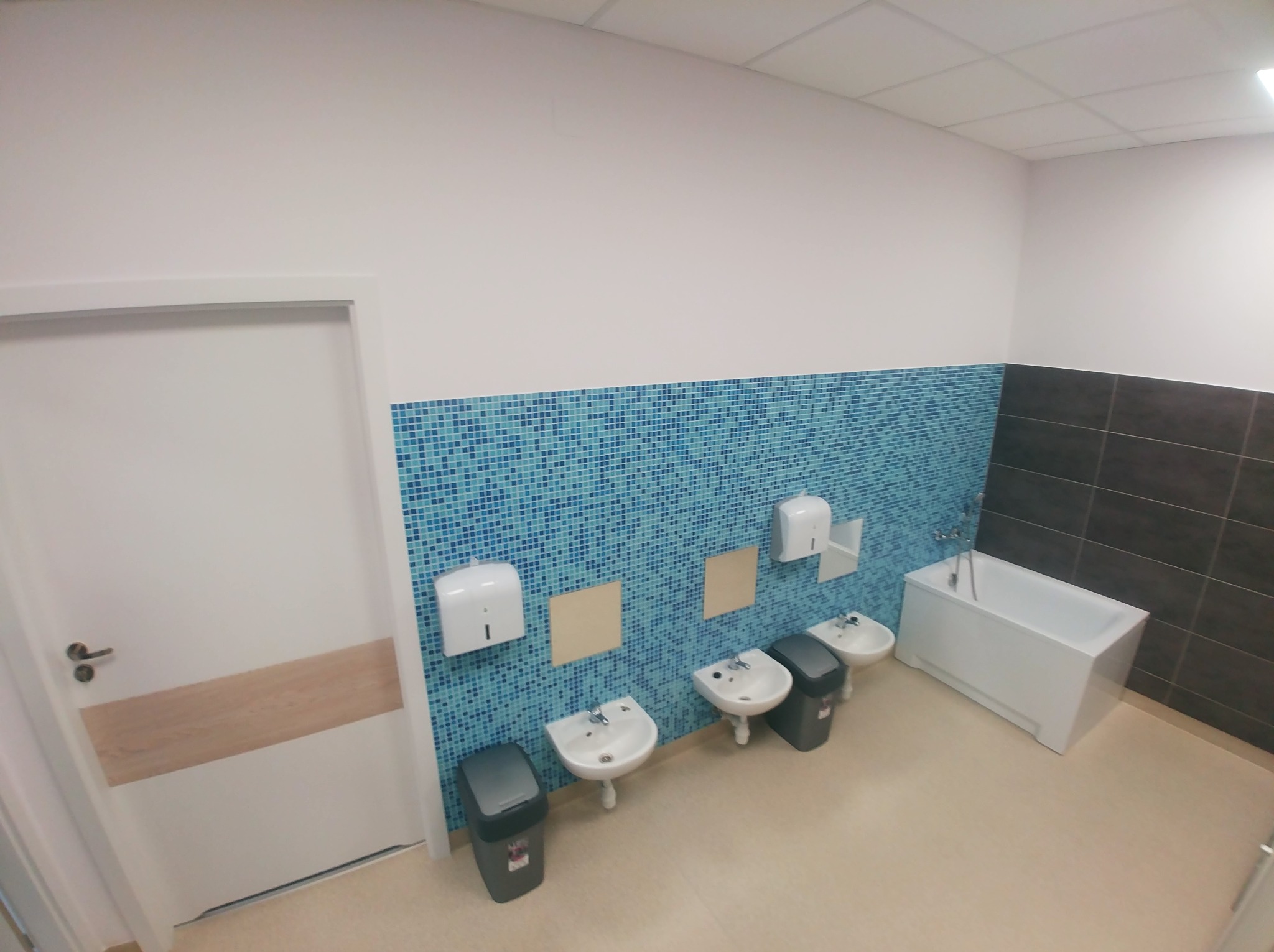 Wnętrze łazienki biurowej z trzema umywalkami z bateriami, pojemnikami na śmieci, dozownikami mydła i ręczników, wykończone mozaiką w odcieniach niebieskiego i brązowymi płytkami, z białą wanną...