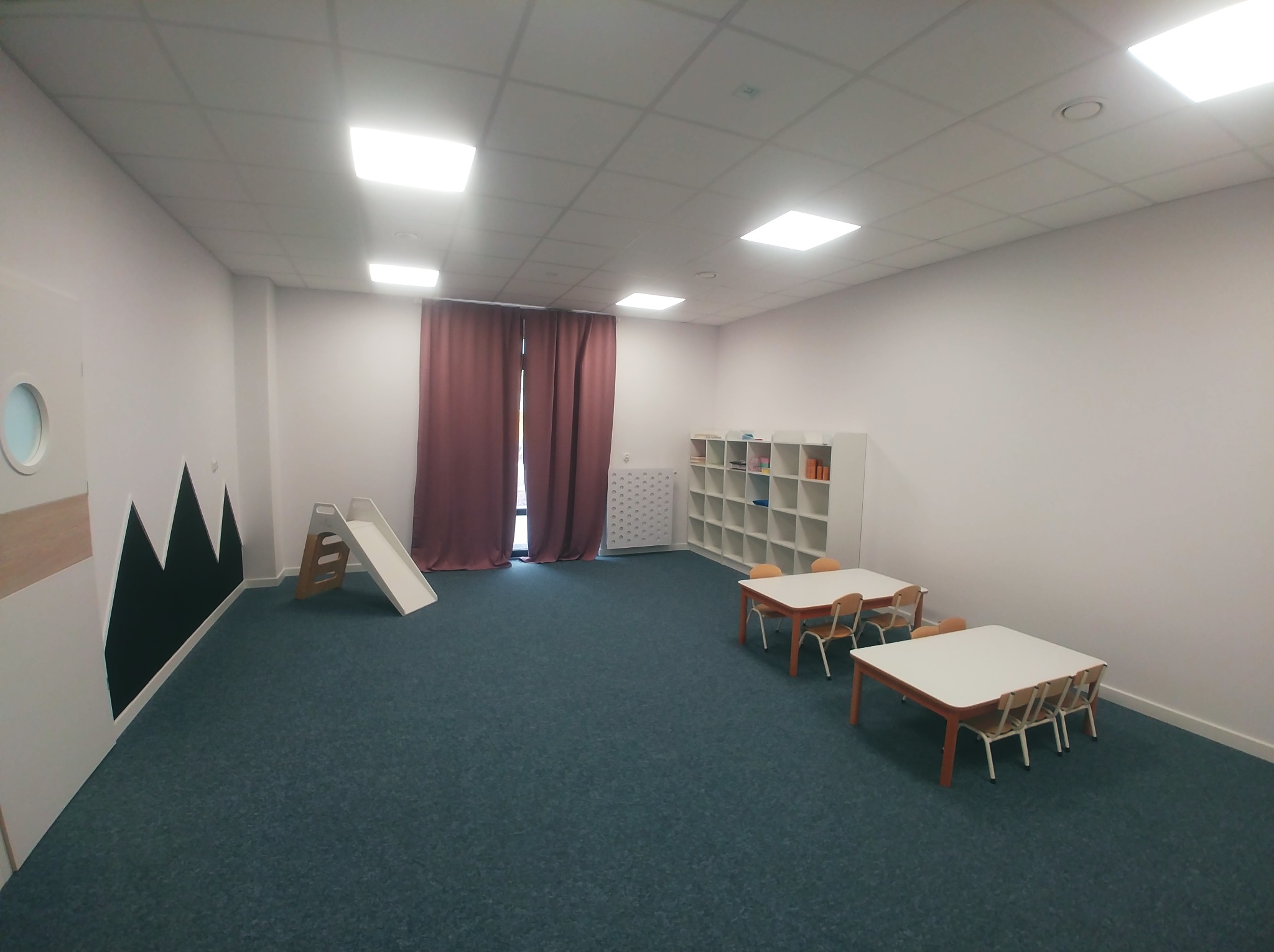 Jasne wnętrze sali zabaw z elementami wyposażenia: stoliki z krzesłami, regał na zabawki, mała zjeżdżalnia oraz dekoracja ścienna w postaci czarnych gór na białym tle.