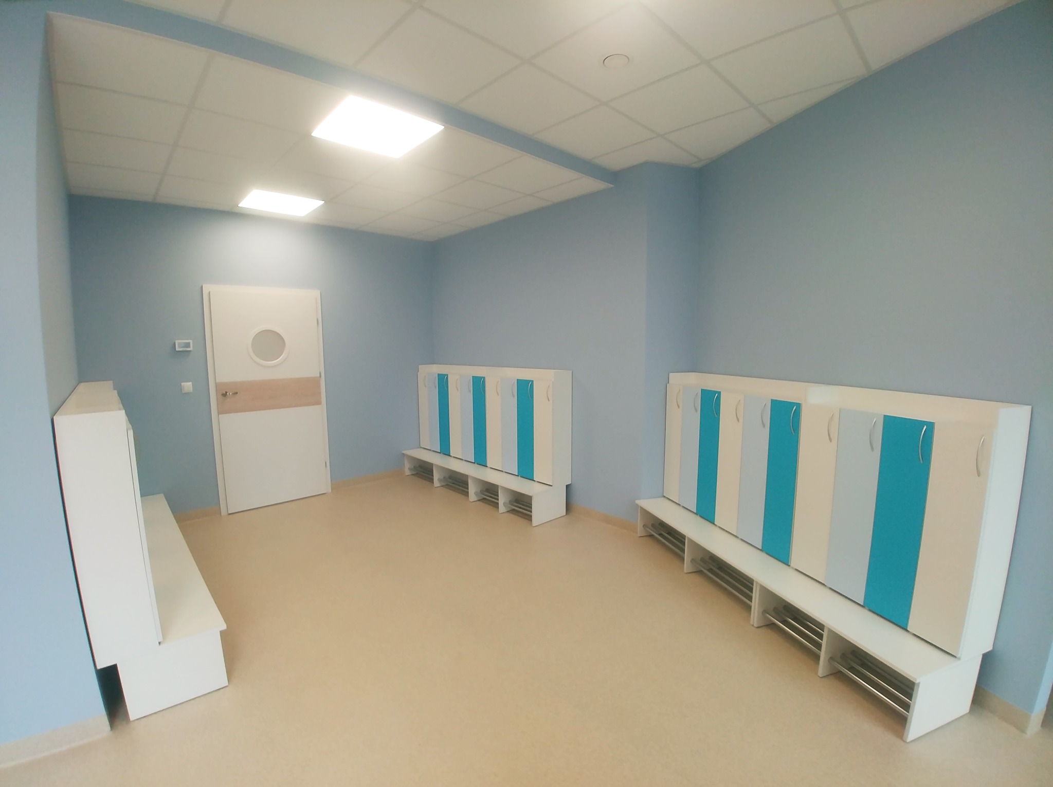 Wyremontowana szatnia w biurze w Warszawie, z jasnoniebieskimi ścianami, białymi szafkami z turkusowymi akcentami i siedziskami, oraz jasną podłogą. Widoczny fragment sufitu podwieszanego...