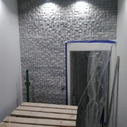 Ściana wykończona dekoracyjnymi panelami 3D w odcieniach szarości, oświetlona dwoma punktowymi lampami sufitowymi, widoczne zabezpieczone folią drzwi i drewniany podest roboczy.