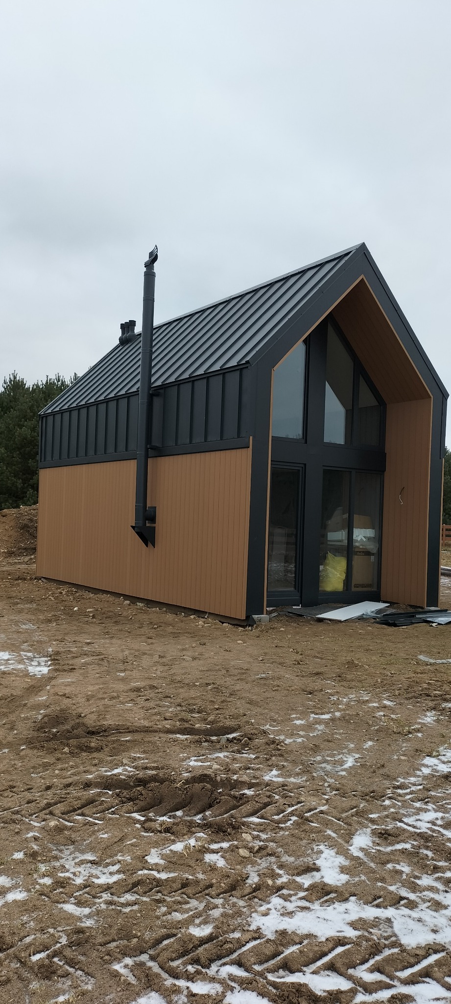 Nowoczesny dom z ciemną, metalową fasadą i drewnianą elewacją boczną, widoczny komin, duże okna i drzwi przesuwne, teren wokół domu częściowo pokryty śniegiem.