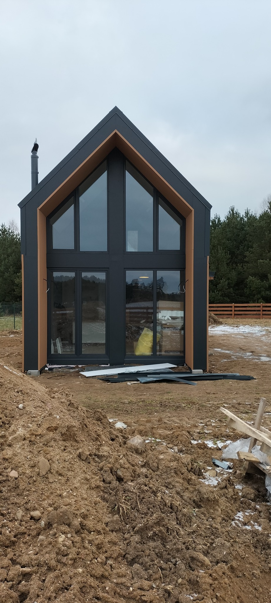 Nowoczesny dom w stylu stodoły z dużymi oknami, czarną elewacją i drewnianymi akcentami, w trakcie budowy, otoczony ziemią i elementami budowlanymi.