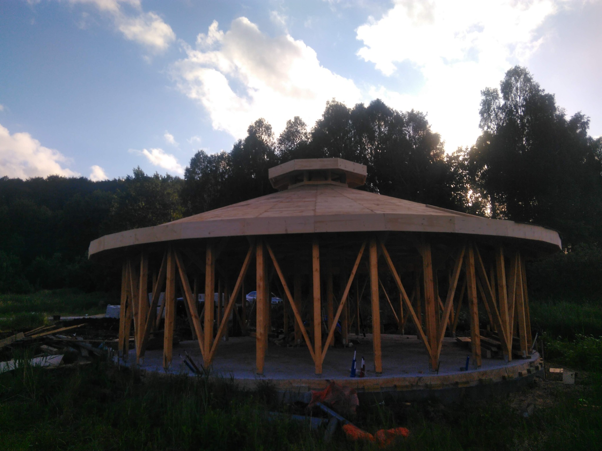 Niedokończona konstrukcja okrągłego domu z drewnianymi słupami i dachem, widoczna na tle lasu i nieba z chmurami.