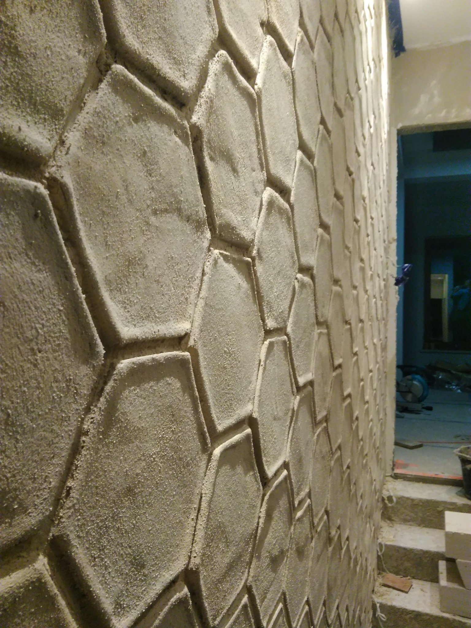 Ściana wewnętrzna wykończona dekoracyjnym tynkiem strukturalnym w kształcie heksagonów, z widocznymi niedoskonałościami i surowym wykończeniem, w tle widoczne schody i sprzęt budowlany.