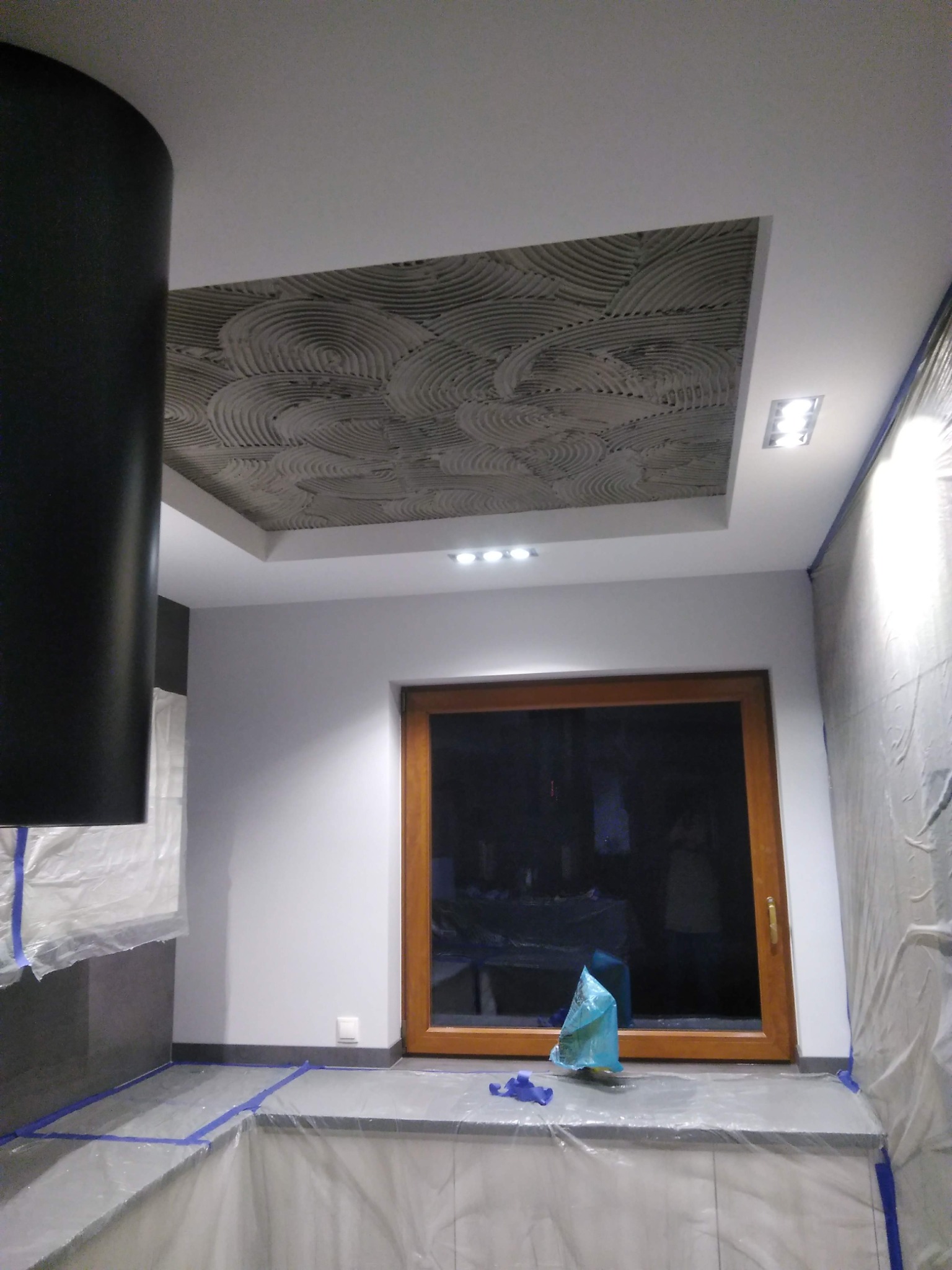 Fragment wnętrza w trakcie prac wykończeniowych, widoczny sufit z dekoracyjną strukturą w kształcie koncentrycznych okręgów, zabezpieczone folią ściany i blat kuchenny, oraz okno w drewnianej ramie.