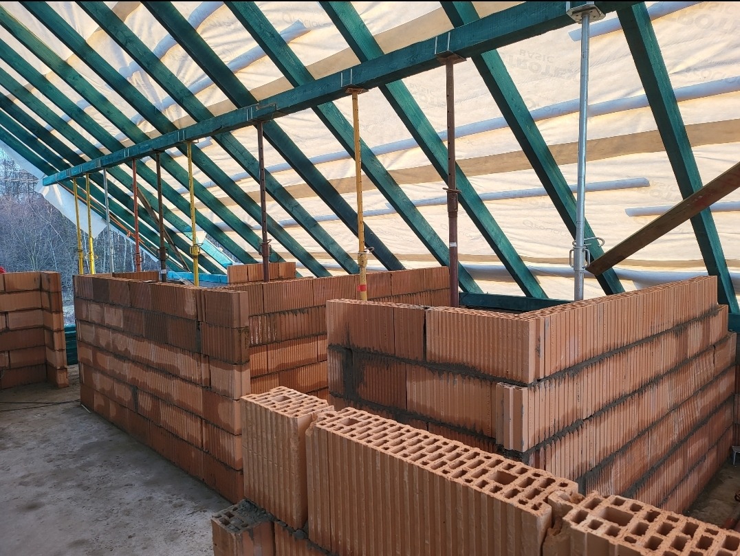 Wznoszenie ścian z czerwonej cegły dziurawki wewnątrz konstrukcji z zielonych belek i białej folii, widoczne podpory stropowe.