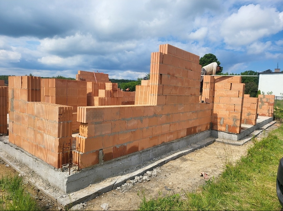 Wznoszenie ścian parteru z czerwonej cegły na fundamencie betonowym, widoczne zbrojenia, w tle zielona roślinność i błękitne niebo z chmurami.