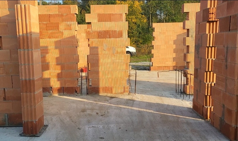 Wznoszone mury z czerwonej ceramiki poryzowanej na fundamencie, widoczne zbrojenia i konstrukcja nośna, w tle zieleń i samochód.