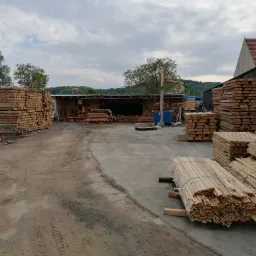 Plac składowania drewna z ułożonymi stosami tarcicy, widoczna hala produkcyjna z maszyną do obróbki drewna w tle, pochmurne niebo.