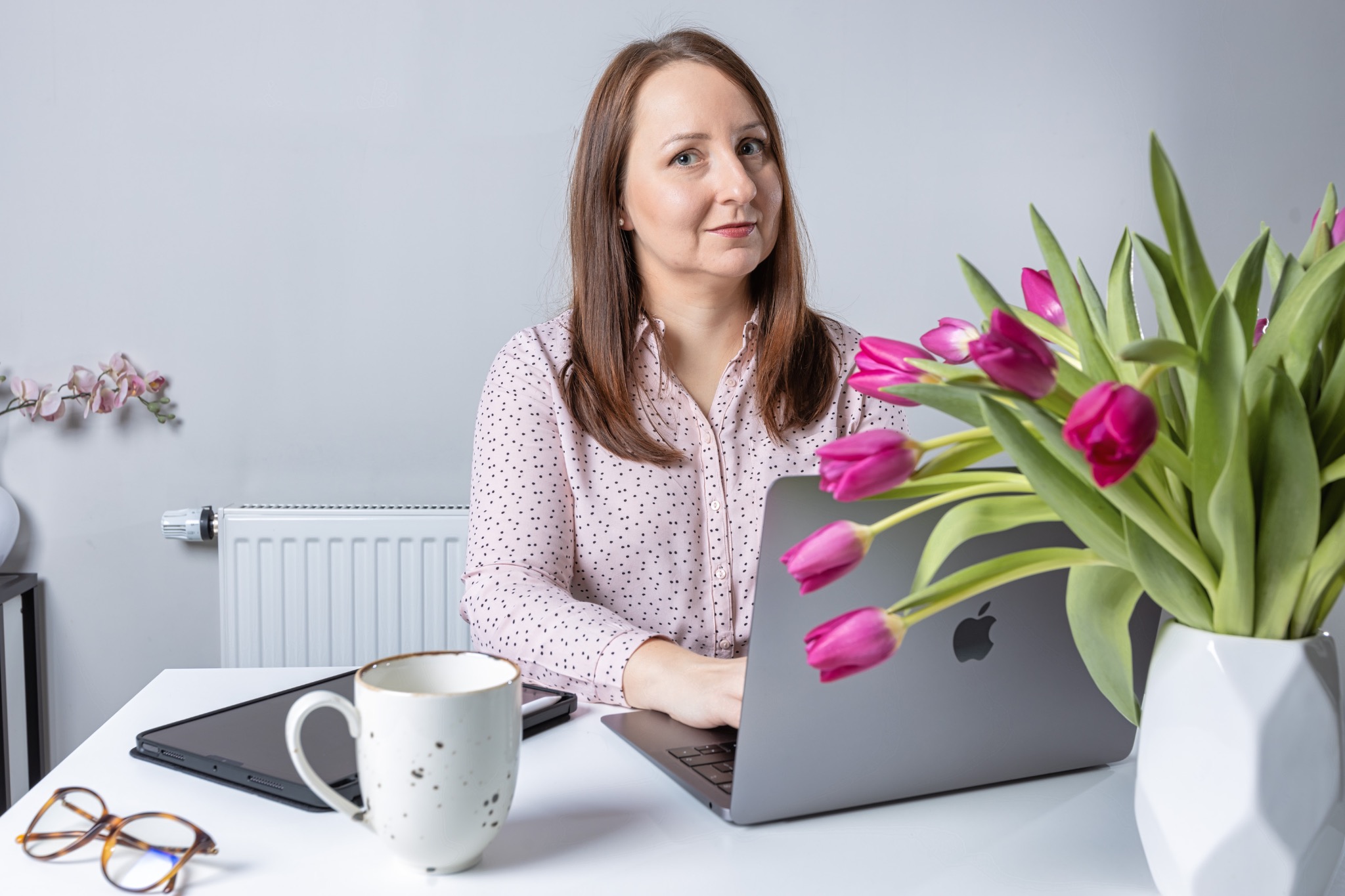 Kobieta w różowej koszuli w kropki pracuje na laptopie Apple; obok wazon z różowymi tulipanami, kubek w plamki i okulary na białym biurku.