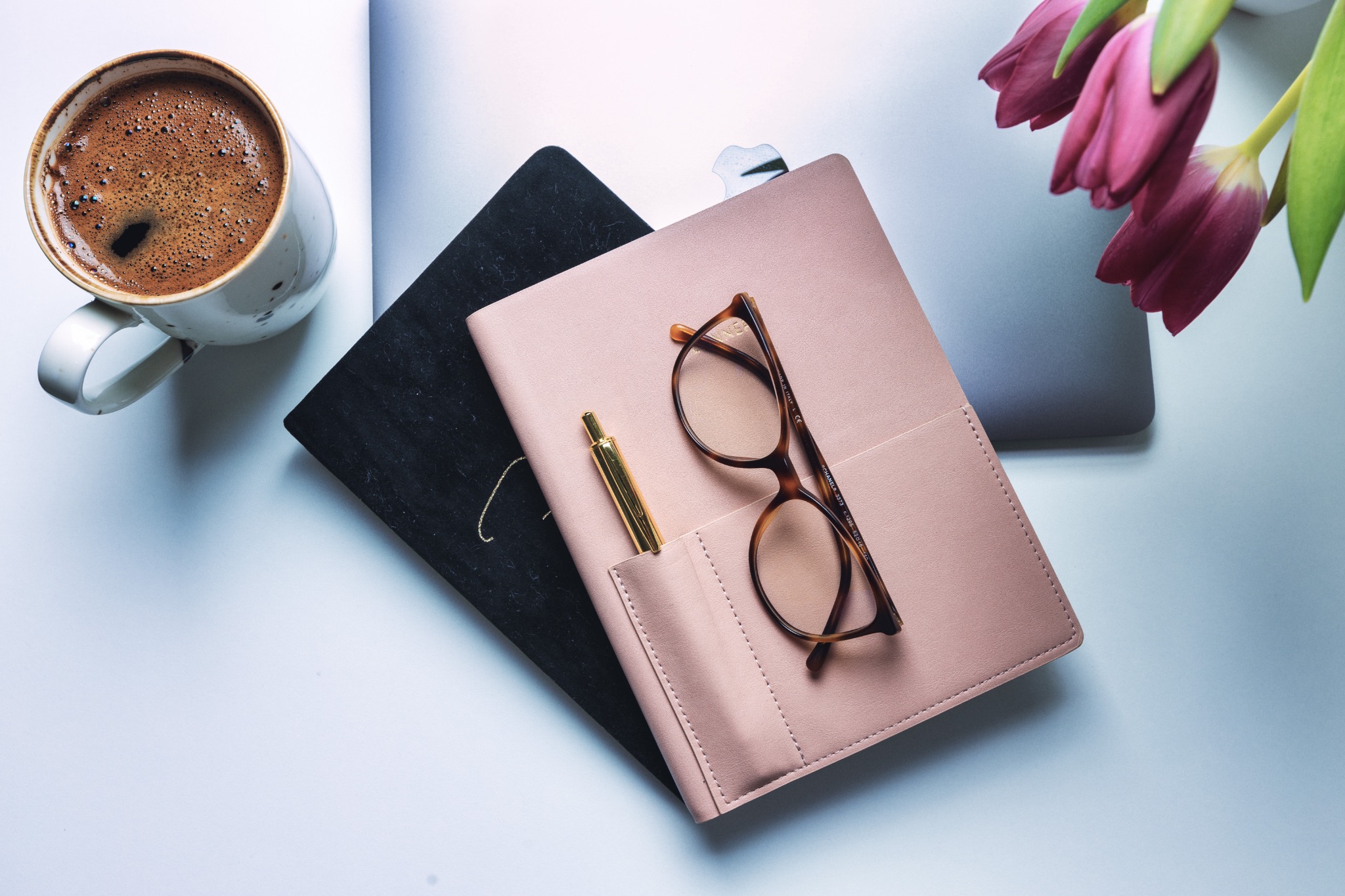Biurko z filiżanką kawy, notatnikami, złotym długopisem, okularami i tulipanami, sugerujące pracę biurową lub pisanie.