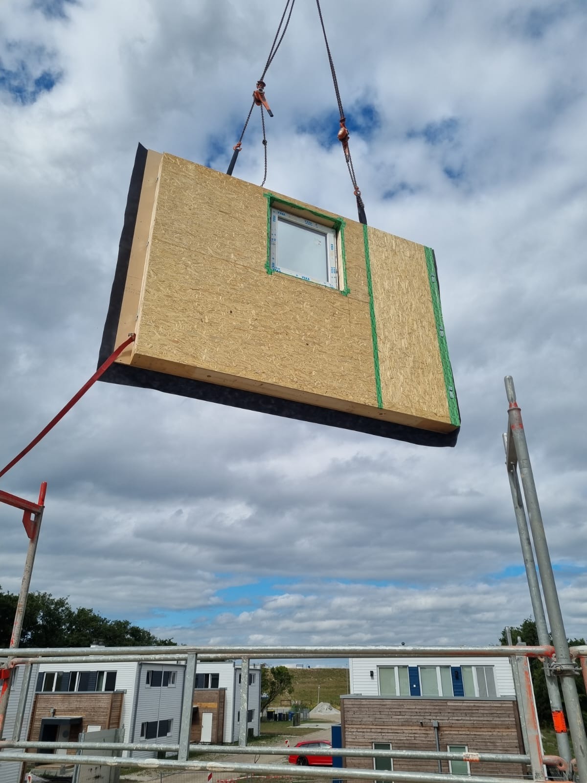 Prefabrykowany element ściany z oknem, podnoszony dźwigiem na placu budowy, widoczne rusztowanie i budynki w tle.