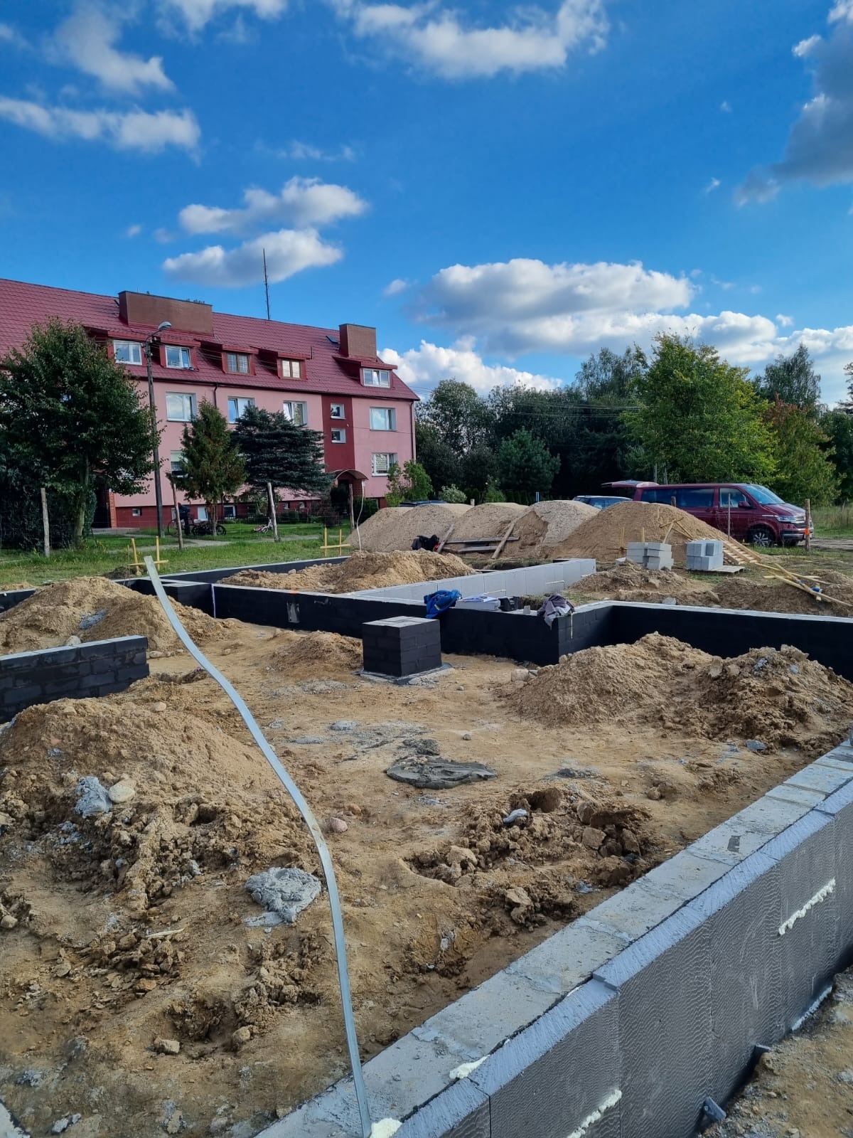 Wykopane fundamenty domu z częściowo wylanym betonem, widoczne bloczki fundamentowe, piasek, budynek mieszkalny w tle i błękitne niebo z chmurami.