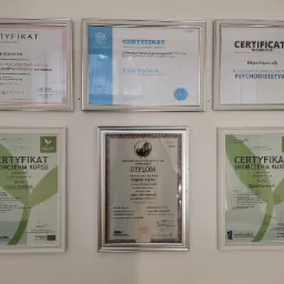 Kolekcja dyplomów i certyfikatów w srebrnych ramkach, wiszących na białej ścianie, potwierdzających kwalifikacje zawodowe Edyty Szymanik w zakresie dietetyki i pokrewnych dziedzin.