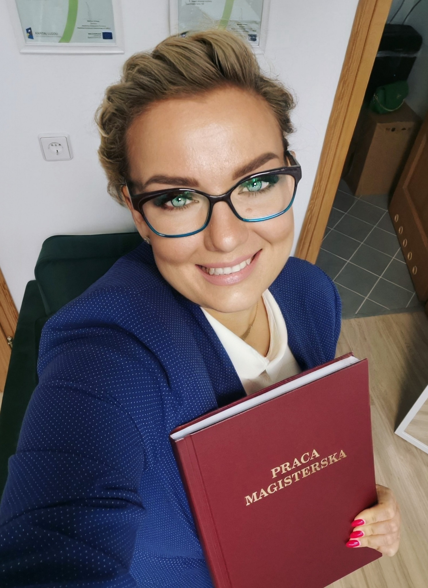 Uśmiechnięta dietetyczka w okularach, trzymająca w dłoniach pracę magisterską z widocznym napisem 'Praca Magisterska', ubrana w granatową marynarkę w białe kropki.