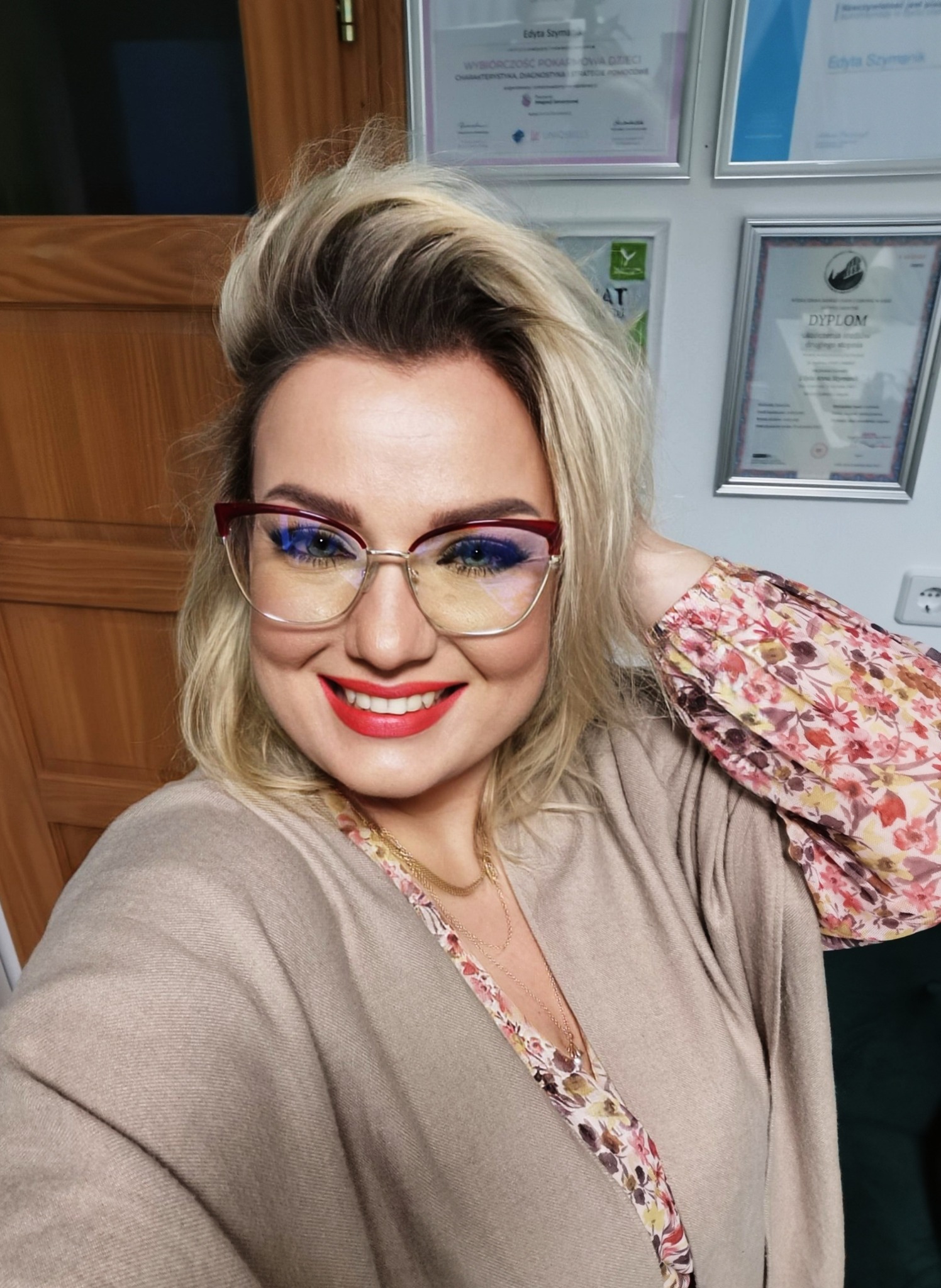Uśmiechnięta kobieta w okularach z czerwoną oprawką, na ścianie dyplomy i certyfikaty w ramkach.