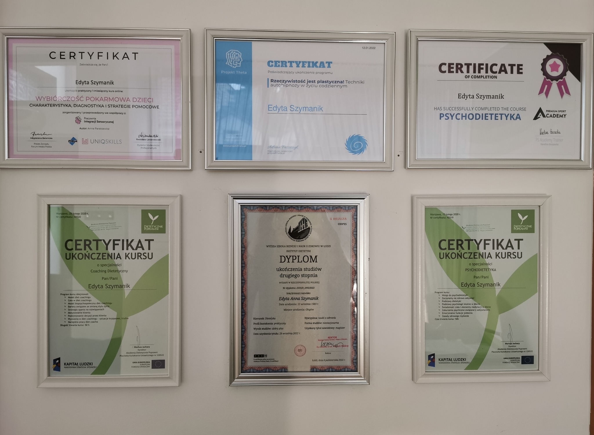 Kolekcja dyplomów i certyfikatów w srebrnych ramkach, wiszących na białej ścianie, potwierdzających kwalifikacje zawodowe Edyty Szymanik w zakresie dietetyki i pokrewnych dziedzin.