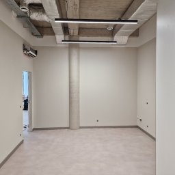 KG Koncept Kacper Gucz - Jasne, przestronne wnętrze biurowe z kolumną, surowym betonowym sufitem z oświetleniem liniowym i klimatyzacją. Widoczne ślady po pracach wykończeniowych na ścianach.