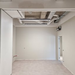 KG Koncept Kacper Gucz - Wnętrze po remoncie: gładkie, jasne ściany, betonowy sufit z instalacjami, nowoczesne oświetlenie liniowe. Widoczne drzwi harmonijkowe i fragment sąsiedniego pomieszczenia.