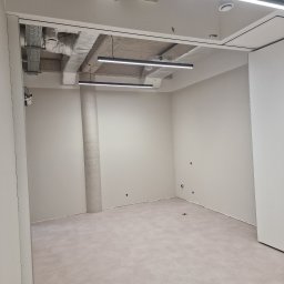 KG Koncept Kacper Gucz - Jasne, puste pomieszczenie biurowe po remoncie, z widocznymi elementami instalacji pod sufitem i kolumną. Ściany pomalowane na jasny odcień, podłoga wyłożona panelami.