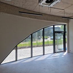 KG Koncept Kacper Gucz - Wnętrze biura po remoncie: gładkie ściany w odcieniach szarości, duże okno z widokiem na zewnątrz, surowy betonowy sufit z nowoczesnym oświetleniem.