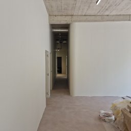 KG Koncept Kacper Gucz - Wnętrze w trakcie wykańczania: gładkie, białe ściany, surowy betonowy sufit, widoczne materiały budowlane. Perspektywa długa, wąska, z widokiem na dalsze pomieszczenia.