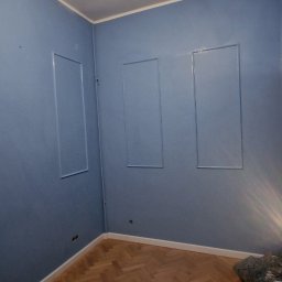 KG Koncept Kacper Gucz - Błękitne ściany z dekoracyjnymi ramkami i parkietem w jodełkę. Widoczna żarówka. Wykończone wnętrze po malowaniu, gotowe do umeblowania.