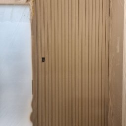 KG Koncept Kacper Gucz - Świeżo pomalowana, beżowa ściana z pionowymi panelami, zabezpieczona folią malarską. Widoczny fragment drzwi z folią i wałek malarski na podłodze.