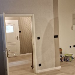 KG Koncept Kacper Gucz - Jasne wnętrze po odświeżeniu: ściany w odcieniach szarości z akcentem beżu, widoczne puszki z farbą, podłoga z paneli. W tle otwarte drzwi do kolejnych pomieszczeń.